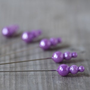 May include: Close-up of several purple decorative pins with a glossy finish. Each pin features a series of three or four rounded beads. The pins are arranged diagonally against a grey wooden surface.