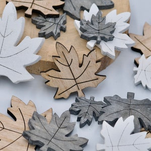 Può includere: Una collezione di ritagli di foglie d'acero in legno in vari colori, tra cui bianco, grigio e tonalità legno naturale. Le foglie sono disposte su uno sfondo chiaro, alcune su una fetta di legno.