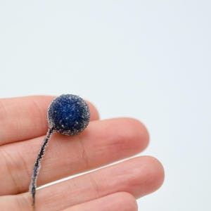 May include: A single, dark blue, frosted berry with a thin stem on a person's hand.