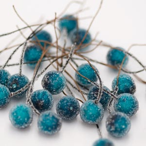 May include: A cluster of teal blue frosted berries on brown wire stems. The berries are arranged in a random pattern and are scattered across a white background.
