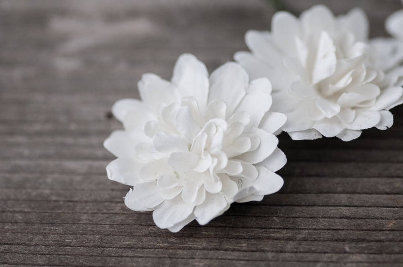 May include: Three white paper flowers with delicate, layered petals. The flowers are arranged on a wooden surface.