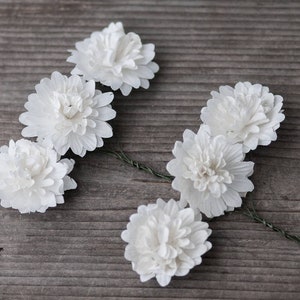 May include: Six white fabric flowers with a green wire stem. The flowers are arranged in a semi-circle on a wooden surface.