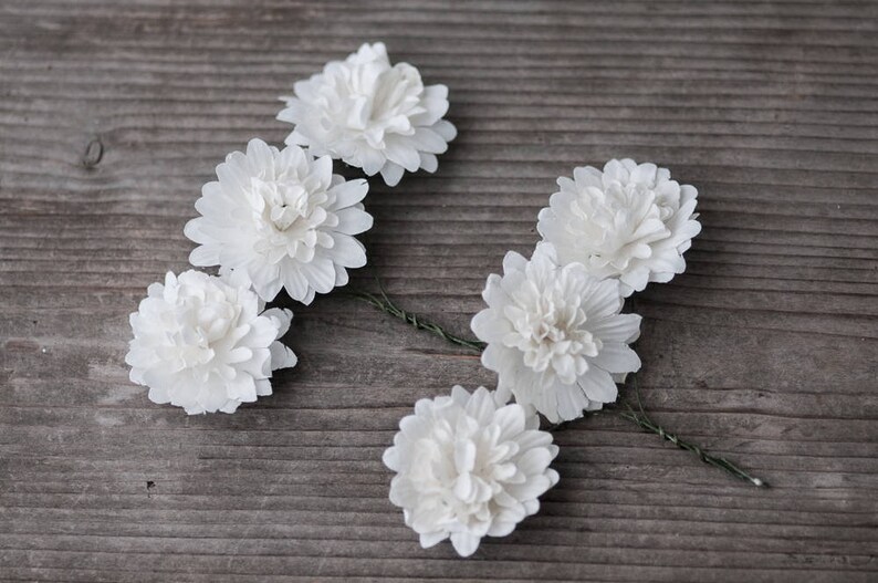 May include: Six white fabric flowers with a green wire stem. The flowers are arranged in a semi-circle on a wooden surface.