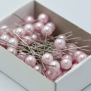 Può includere: Una scatola bianca piena di spilli con testa di perla rosa. Gli spilli sono disposti in modo casuale.