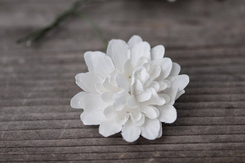May include: A single white paper flower with multiple layers of petals. The flower is on a wood background.