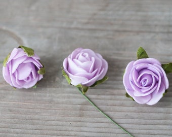 May include: Three lavender paper roses with green stems and leaves. The roses are arranged in a row on a light gray wooden surface.
