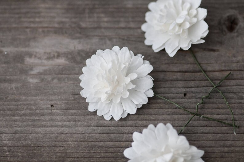 May include: Three white paper flowers with green stems on a wooden surface.