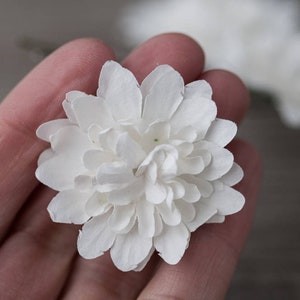 May include: A single white paper flower with multiple layers of petals. The flower is held in a hand.