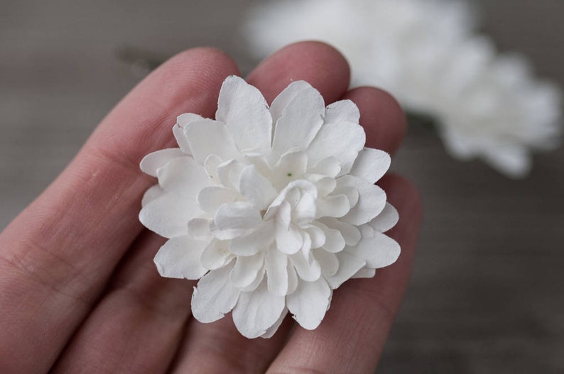 May include: A single white paper flower with multiple layers of petals. The flower is held in a hand.