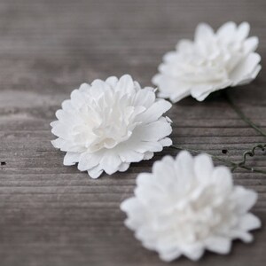 May include: Three white paper flowers with green wire stems on a gray wooden surface.