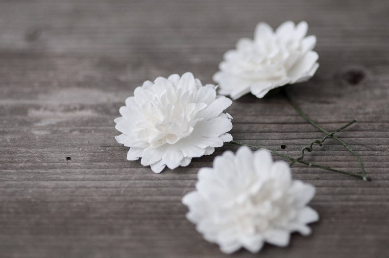 May include: Three white paper flowers with green wire stems on a gray wooden surface.