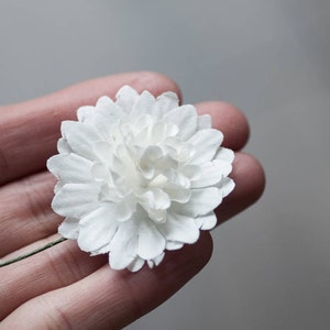 May include: A single white paper flower with a green stem. The flower is made of many small petals and is held in a hand.