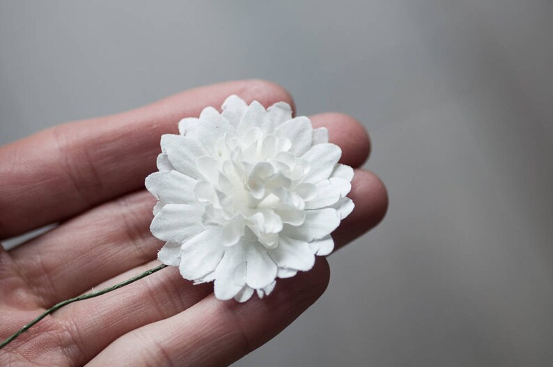 May include: A single white paper flower with a green stem. The flower is made of many small petals and is held in a hand.