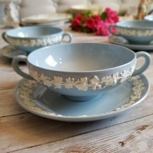 May include: Light blue soup bowls with handles and matching saucers. The bowls and saucers have a white floral relief design around the rims. The bowls are sitting on a light wooden surface.