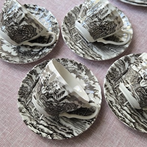 May include: A collection of vintage teacups and saucers. The cups and saucers feature a black and grey landscape pattern on a white background. Each cup has a curved handle and rests on a matching saucer. The set is displayed on a pink surface.