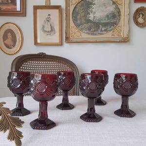 Op de afbeelding: Zes robijnrode wijnglazen met sierlijke bloemen- en geometrische patronen. De glazen hebben een voet en een gestructureerd ontwerp. De glazen zijn gerangschikt op een wit tafelkleed, met antieke ingelijste kunstwerken op de achtergrond.