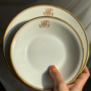 Puede incluir: Dos platos de porcelana blanca con bordes dorados y un monograma dorado. Los platos se sostienen en una mano. Los platos son de diseño clásico, adecuados para cenas formales u ocasiones especiales.