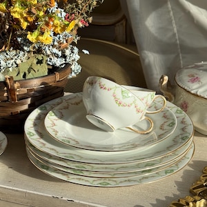 May include: A vintage tea set featuring white porcelain cups and saucers with delicate pink floral and green leaf patterns. The set includes gold trim and handles, with a stack of plates and a teacup resting on top.