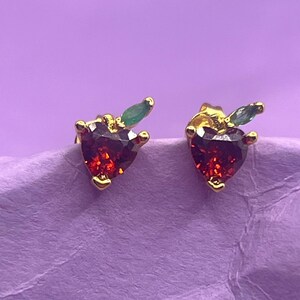 Pode incluir: Um par de brincos de botão dourados. Cada brinco apresenta uma pedra preciosa vermelha escura em forma de coração com um pequeno detalhe de folha verde. Os brincos estão sobre um fundo roxo.