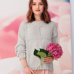 May include: A light gray knitted sweater with a textured pattern. The sweater has a relaxed fit and a boat neck. The model is holding a bouquet of pink hydrangeas.