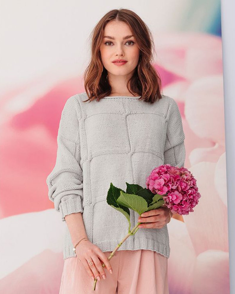 May include: A light gray knitted sweater with a textured pattern. The sweater has a relaxed fit and a boat neck. The model is holding a bouquet of pink hydrangeas.