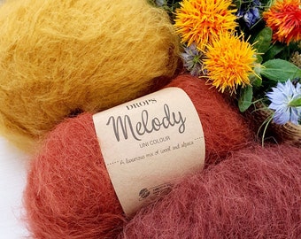 May include: Three balls of yarn in beige, yellow, and red. The yarn is a blend of wool and alpaca. The label reads "DROPS Melody Uni Colour... A luxurious mix of wool and alpaca."