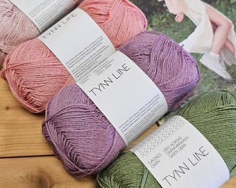 Puede incluir: Primer plano de varios ovillos de hilo Tynn Line en varios colores, incluyendo rosa, morado y verde. Cada ovillo tiene una etiqueta blanca con el nombre de la marca. El hilo está dispuesto sobre una superficie de madera.