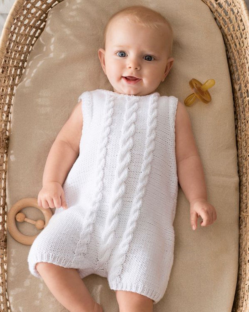 May include: A white knitted baby romper with a cable knit pattern. The romper is sleeveless and has a round neckline. The baby is lying on its back in a wicker basket.