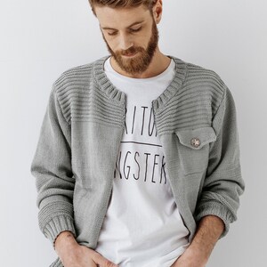 May include: A gray knitted cardigan with a buttoned pocket on the left side. The cardigan has a ribbed texture and a slightly open collar.
