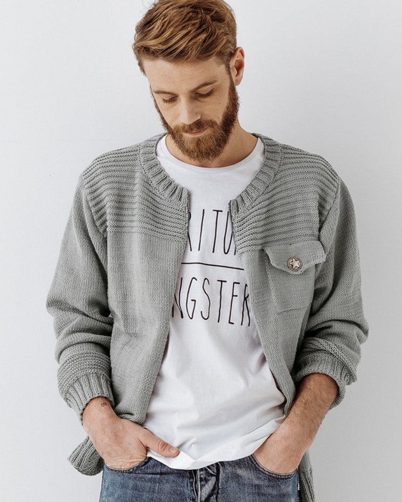 May include: A gray knitted cardigan with a buttoned pocket on the left side. The cardigan has a ribbed texture and a slightly open collar.