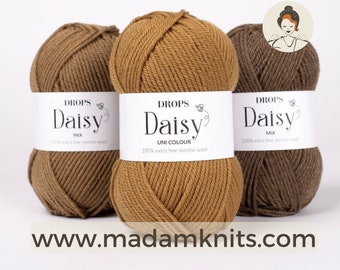 Puede incluir: Tres ovillos de lana en diferentes tonos de marrón. La lana está etiquetada como "DROPS Daisy" y está hecha de 100% lana merino extra fina. La etiqueta también indica que un ovillo es "Mix" y otro es "Uni Colour".