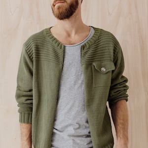 May include: A green knitted cardigan with a buttoned pocket on the left side. The cardigan has a ribbed collar and cuffs.
