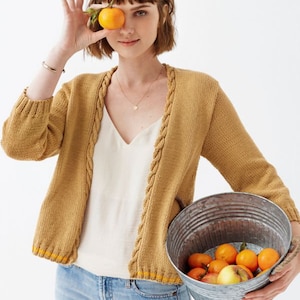 May include: A woman wearing a mustard yellow cardigan with a cable knit detail and a white tank top. She is holding a bucket of oranges and an orange in her hand.