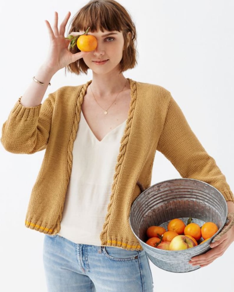 May include: A woman wearing a mustard yellow cardigan with a cable knit detail and a white tank top. She is holding a bucket of oranges and an orange in her hand.