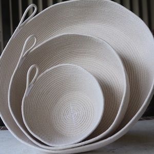 May include: Three white cotton rope bowls of varying sizes, nested together. The bowls have a woven texture and are made of natural materials.