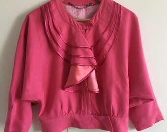 May include: A pink silk blouse with a large ruffled collar and a button closure. The collar is made of multiple layers of pink silk and has a slightly darker pink hue at the bottom.
