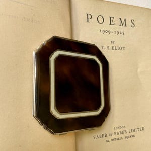 May include: A vintage tortoiseshell compact with a silver frame, resting on the open pages of a book titled "Poems" by T.S. Eliot. The compact has a geometric design with a central dark brown area surrounded by a white and light brown border.