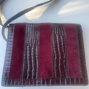 May include: A burgundy and brown shoulder bag with a black strap. The bag features a combination of textures, with suede panels and faux crocodile leather accents. The bag is rectangular in shape.