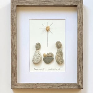 May include: A framed pebble art piece featuring a nativity scene made of smooth gray and tan stones. Above the figures is a star formed by a single stone and thin black lines. The text "Emmanuel... God with us" is written in black ink below.