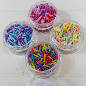 May include: Four small jars filled with colorful sprinkles. The jars are arranged in a circle with a larger jar in the center that has a white label with the text "Sprinkle Company".