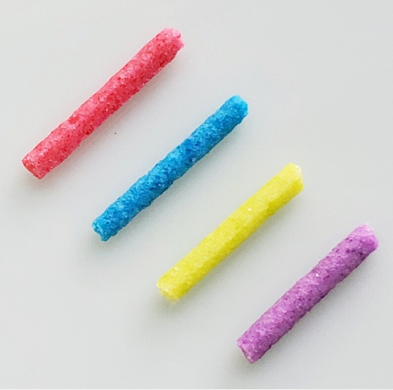 Puede incluir: Cuatro palitos texturizados de colores, rosa, azul, amarillo y morado, dispuestos en fila sobre una superficie blanca.
