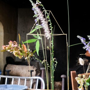 Peut inclure: Une structure géométrique en fil d'or contenant un arrangement de fleurs fraîches et de verdure. L'arrangement comprend un feuillage violet, blanc et vert, posé sur une table en bois. L'arrière-plan présente une cheminée et un décor rustique.