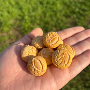 May include: A handful of small, round, yellow-orange pendants with leaf-like designs. The pendants are held in an open palm against a blurred green background, suggesting an outdoor setting. The pendants appear to be handmade.