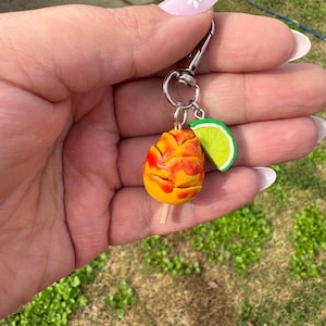 May include: A keychain featuring a miniature mango on a stick, with red and yellow coloring, and a green lime slice charm. The keychain has a silver clasp and ring.