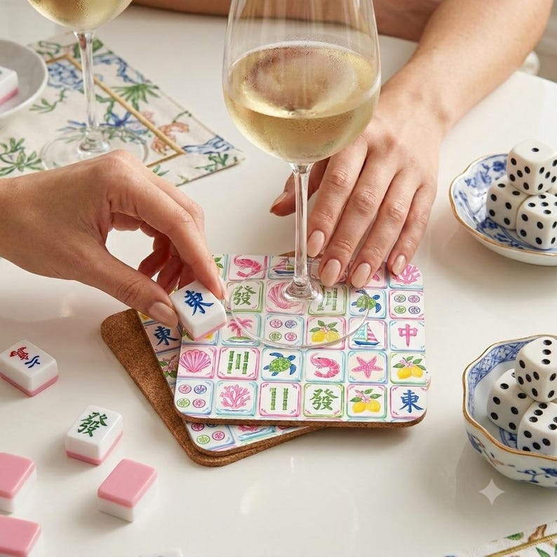 May include: A square cork-backed coaster featuring a grid of colorful illustrations including mahjong tiles, sea life, and fruit. The coaster sits under a wine glass, with loose mahjong tiles and white dice with black dots nearby on a table.