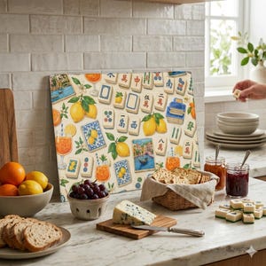 May include: A rectangular cutting board with a colorful design featuring lemons, cocktails, and Mahjong tiles. The board is surrounded by food items like fruit, cheese, crackers, and jam jars, creating a vibrant kitchen scene.