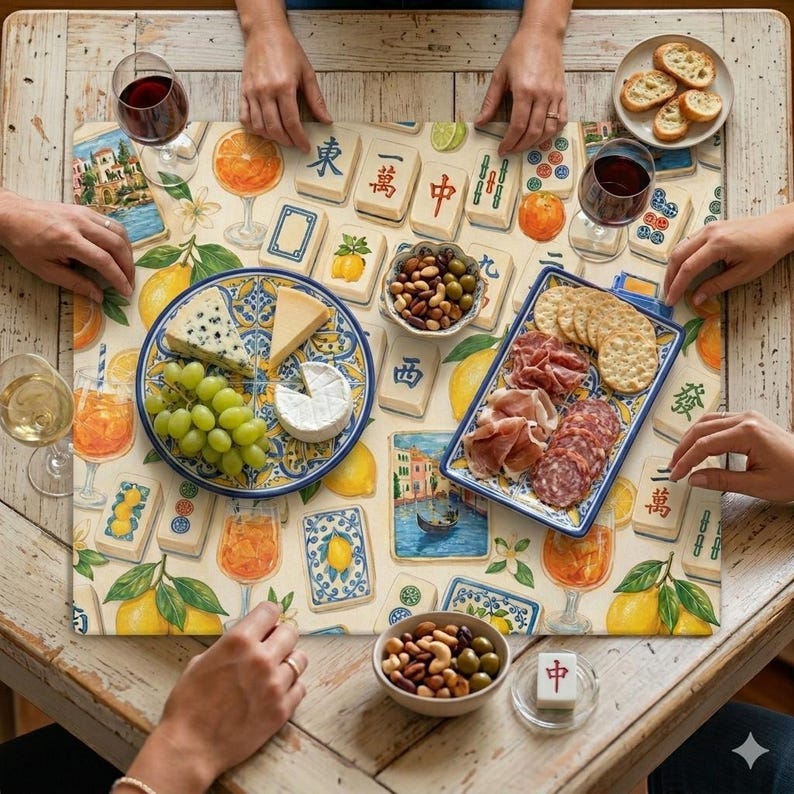 May include: A table setting with a cream-colored placemat featuring lemons, oranges, and Mahjong tiles. The table is set with plates of cheese, grapes, crackers, and meats. Wine glasses and small bowls of nuts and olives are also present.