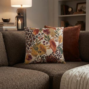 May include: A square throw pillow featuring a cream-coloured background with a printed pattern of autumn leaves, berries, and leopard print spots. The pillow is positioned next to a solid dark brown velvet throw pillow on a textured brown sofa.