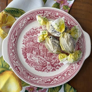 May include: A white and red decorative plate with handles, featuring a scenic design. Several wrapped candies are on the plate. The plate rests on a floral patterned cloth with lemons and pink flowers.