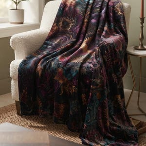 Puede incluir: Una manta de felpa con un estampado botánico oscuro y evocador, con motivos florales y de follaje en tonos púrpura intenso, azul verdoso y dorado. La tela tiene una textura aterciopelada y está colocada sobre un sillón blanco texturizado.